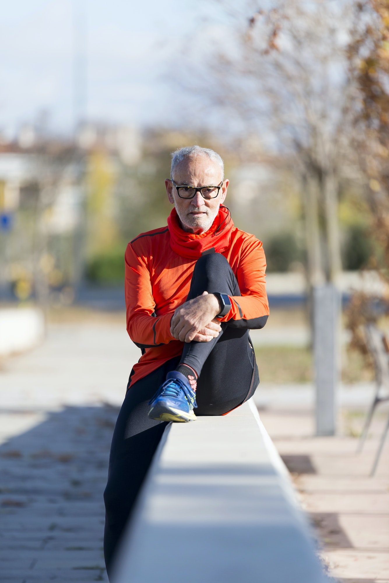 Front view of a senior man wearing sportswear and eyeglasses sitting on a fence