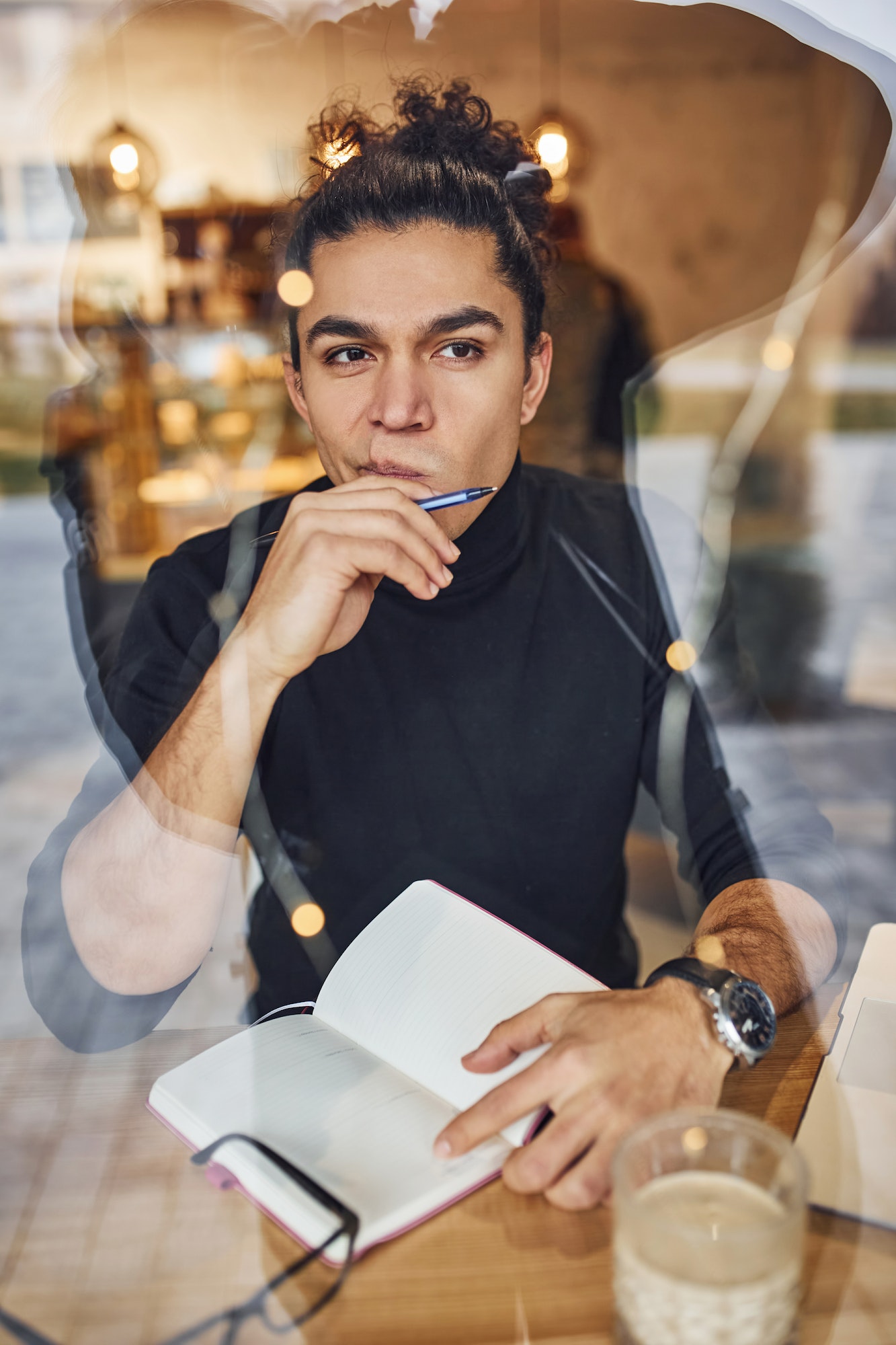 Young male writer with black curly hair sitting indoors in cafe with pen and notepad